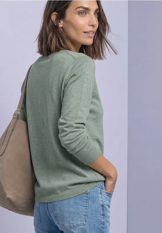 Frau im Profil mit grünem Pullover, blauen Jeans und beiger Tasche über der Schulter