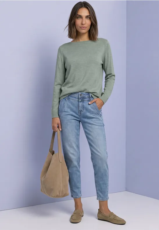 Frau in grünem Pullover, Jeans und braunen Slippern trägt eine beige Tasche vor lilafarbenem Hintergrund.