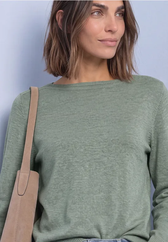 Frau mit schulterlangem braunem Haar, trägt grünen Pullover und beige Umhängetasche vor hellem Hintergrund.