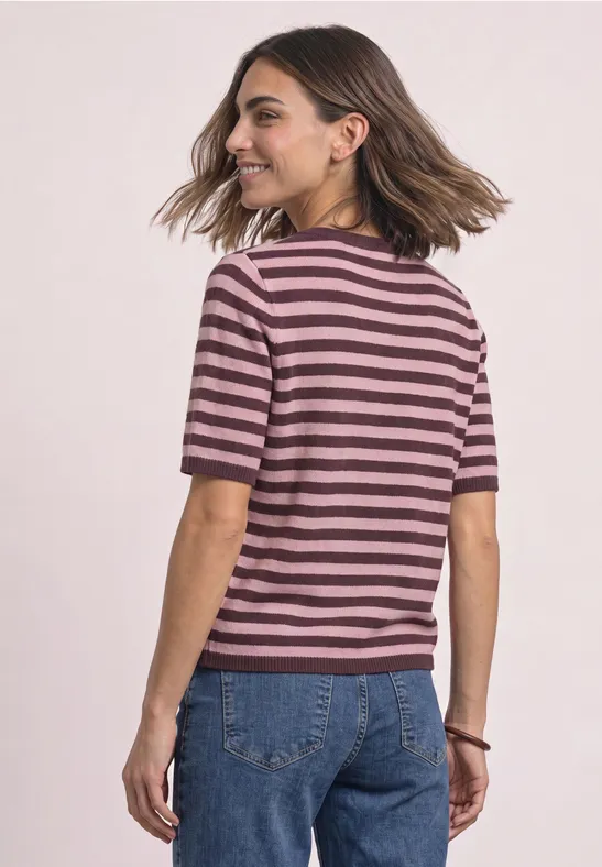 Frau trägt rosa-bordeaux gestreiftes T-Shirt und blaue Jeans, dreht sich lächelnd zur Seite.
