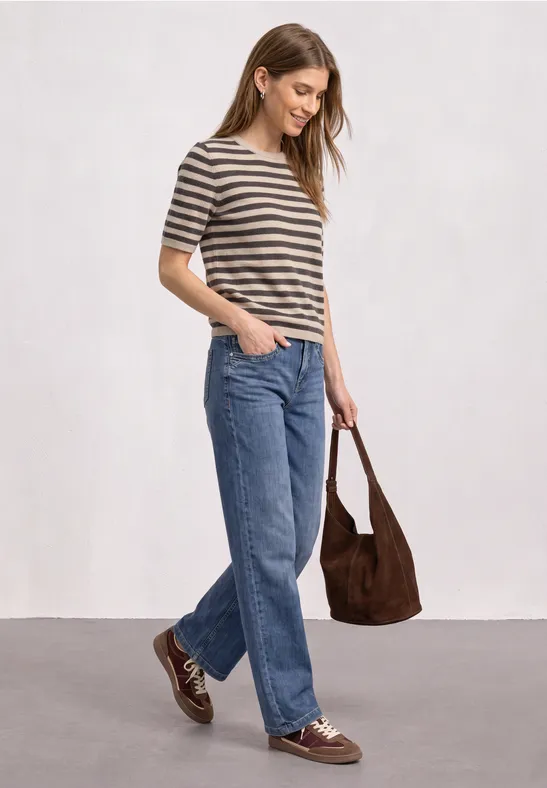 Frau trägt gestreiftes T-Shirt, weite Jeans, braune Schuhe und eine passende Tasche auf hellem Hintergrund.