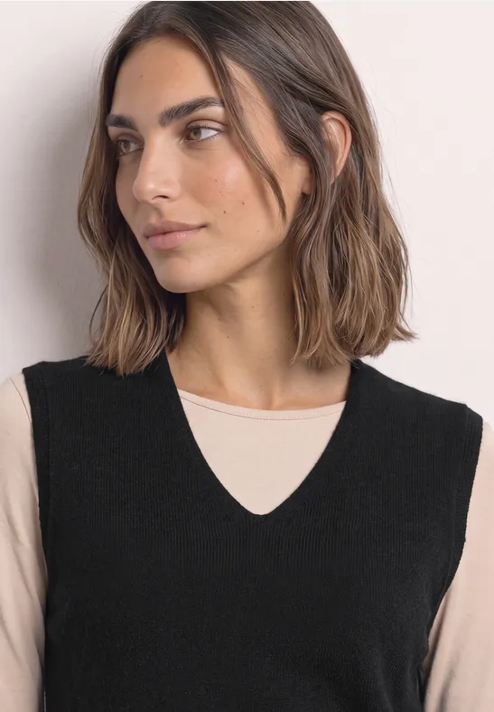 Vrouw in zwart mouwloos vest met V-hals over beige top, zijwaarts kijkend.