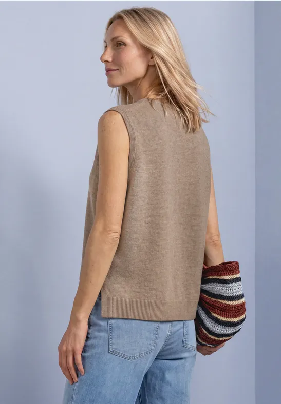 Eine blonde Frau trägt eine beige, ärmellose Strickweste, blaue Jeans und hält eine bunte, gestreifte Tasche.
