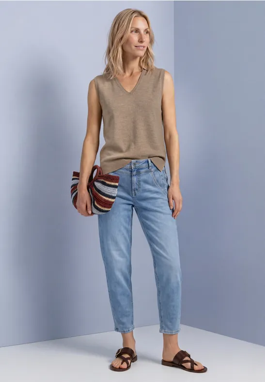 Frau in ärmellosem, beigefarbenem V-Pullover, heller Jeans und schwarzen Sandalen, mit gestreifter Tasche.