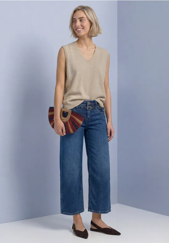 Frau vor hellviolettem Hintergrund trägt beiges ärmelloses Top, weite Cropped-Jeans, flache Schuhe und runde Tasche.