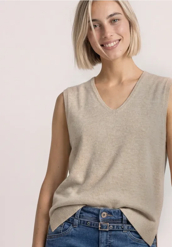 Frau trägt ärmellosen, beigefarbenen V-Ausschnitt-Strickpullunder zu blauen Jeans.