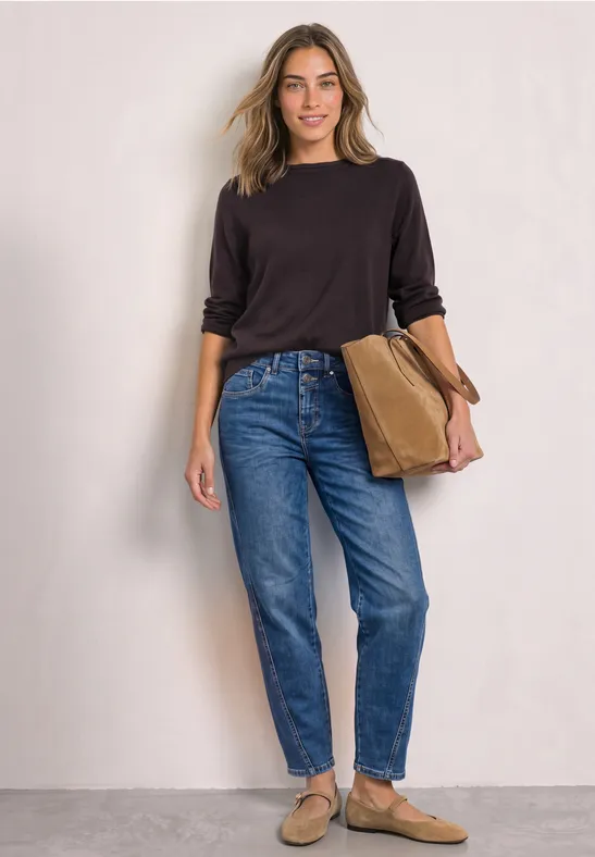 Femme en haut noir, jean bleu et mules beiges, souriante devant un mur neutre, tenant un sac marron.