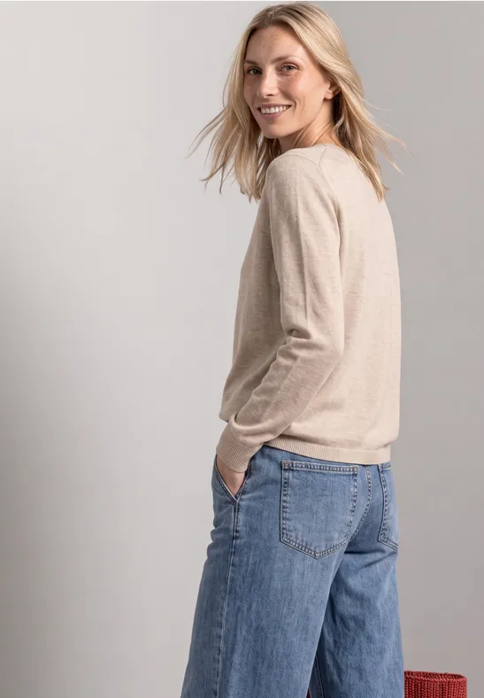 Frau mit beigem Pullover und weiten Jeans, sieht lächelnd über die Schulter.