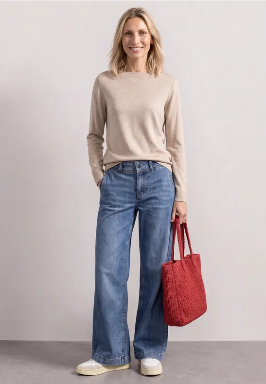 Eine Frau in hellem Pullover und weiten Jeans trägt eine rote Tasche und weiße Sneaker.