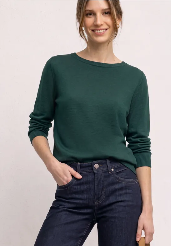Frau trägt dunkelgrünen Pullover und Jeans, lächelt, Hand in Tasche vor hellem Hintergrund.