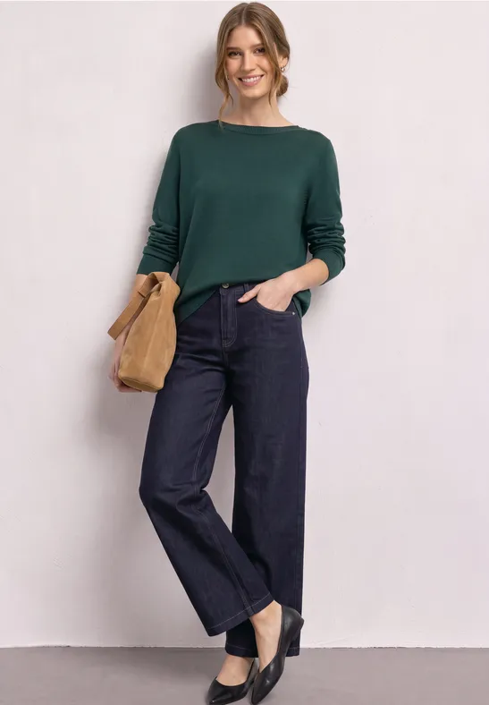 Eine lächelnde Frau trägt einen grünen Pullover, weite Jeans, schwarze Schuhe und hält eine hellbraune Clutch.