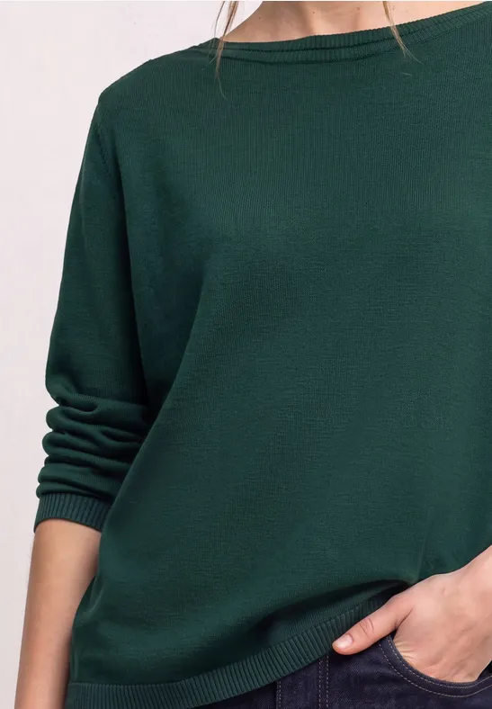 Person trägt locker geschnittenen dunkelgrünen Pullover mit hochgekrempelten Ärmeln, Hand in Tasche.