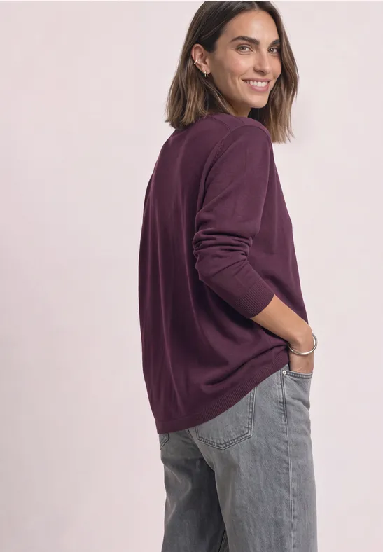 Frau im violetten Pullover und grauen Jeans blickt lächelnd über die Schulter.