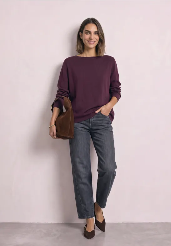 Frau in violettem Pullover, grauen Jeans und dunklen Slippern, mit brauner Tasche vor hellem Hintergrund.