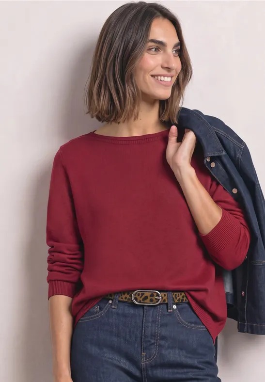 Eine lächelnde Frau in rotem Pullover hält lässig eine blaue Jacke über der Schulter.