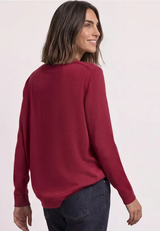 Frau in bordeauxrotem Pullover und Jeans, lächelt über die Schulter.