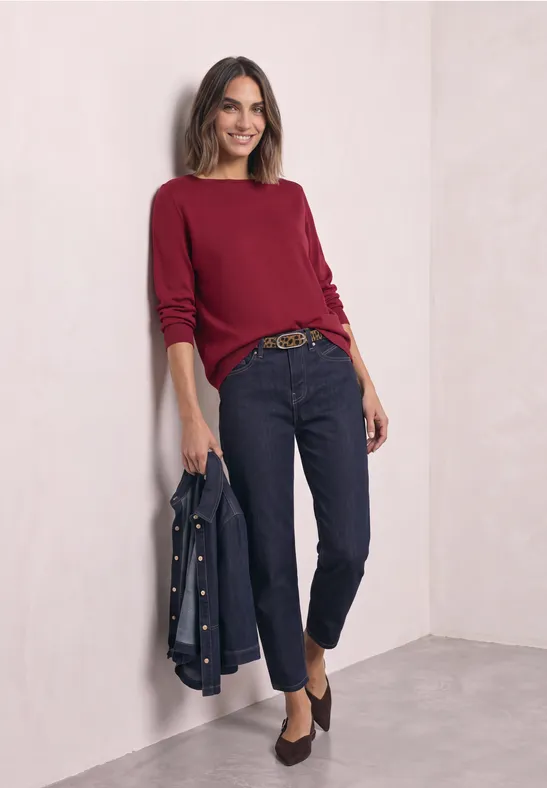 Frau lehnt lächelnd an heller Wand, trägt roten Pullover, dunkle Jeans, Gürtel und hält Jeansjacke