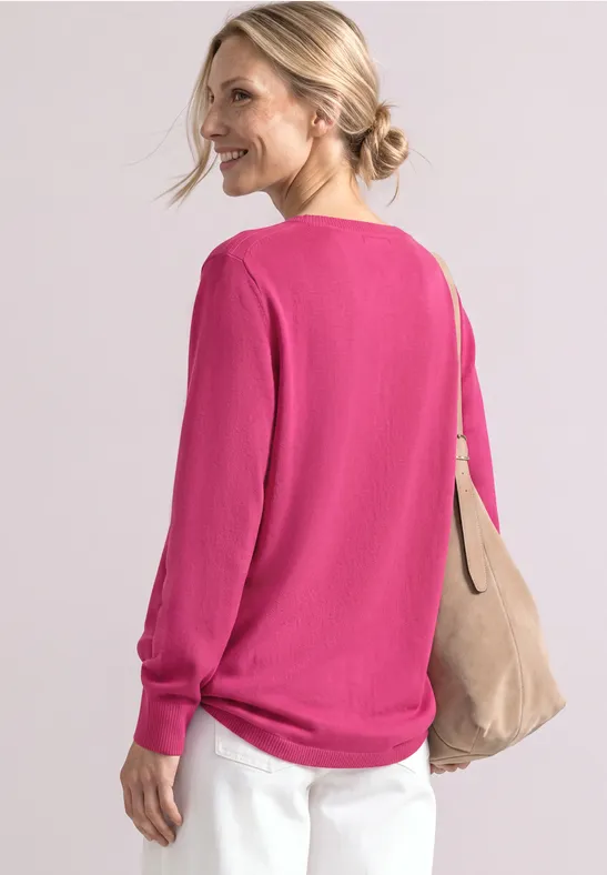 Frau im Profil trägt pinken Pullover, weiße Hose und eine beige Schultertasche.