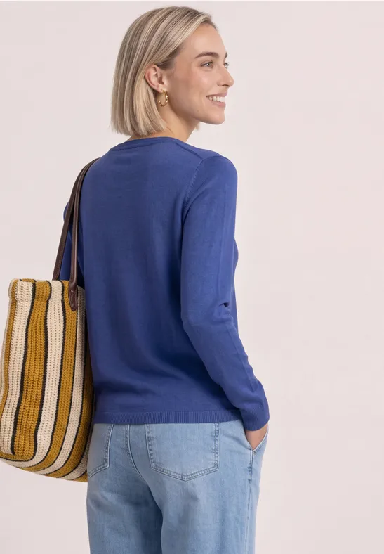 Frau in blauem Pullover und Jeans mit gestreifter Tasche vor hellem Hintergrund.