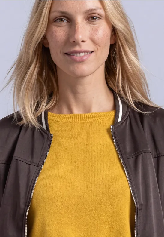 Eine lächelnde Frau mit hellblonden Haaren trägt einen senfgelben Pullover und eine dunkle Jacke vor neutralem Hintergrund.
