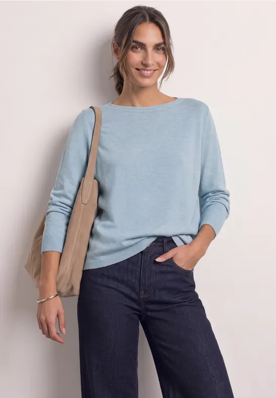 Een glimlachende vrouw in lichtblauwe trui en donkere jeans, met een schoudertas voor een lichte achtergrond.