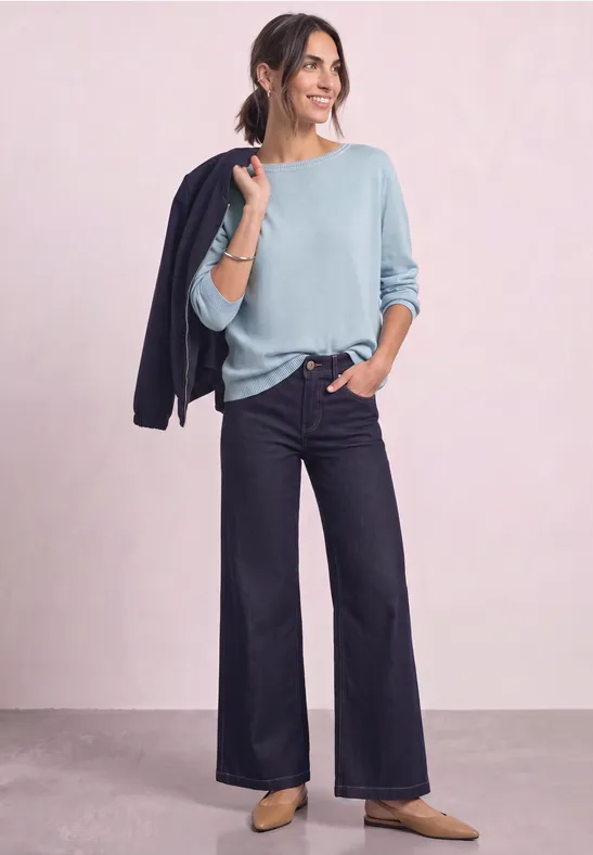 Vrouw in lichtblauwe trui en wijde jeans, nonchalant met een donker jasje over de schouder.