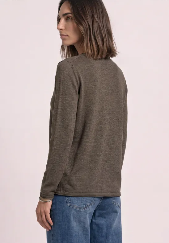 Vrouw in een bruin shirt met lange mouwen en blauwe jeans, schuin van achter gefotografeerd tegen lichte achtergrond.
