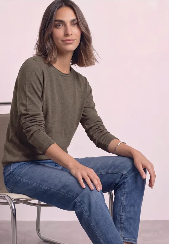 Een vrouw met bruin haar zit ontspannen op een stoel, draagt een groene trui en een blauwe jeans.