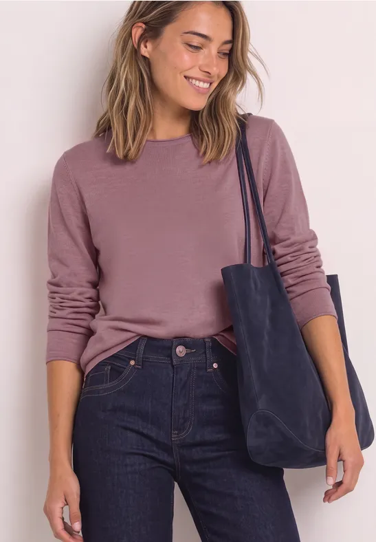 Frau in rosa Pullover und dunklen Jeans trägt eine blaue Tasche und lächelt.