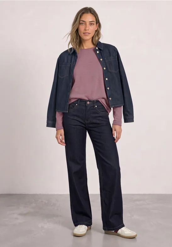 Frau trägt rosa Pullover, weite dunkelblaue Jeans, Jeansjacke auf den Schultern und weiße Sneaker.