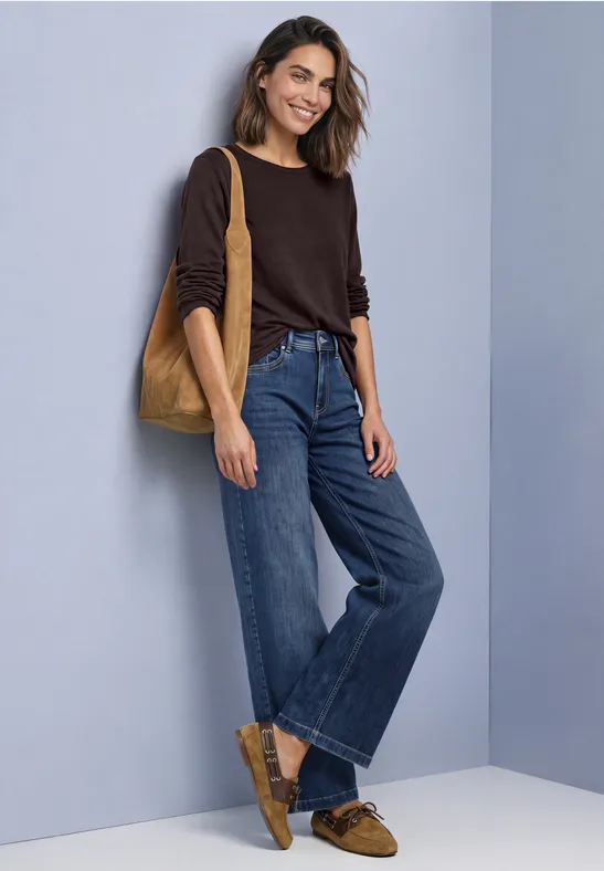 Lächelnde Frau in braunem Pullover, weiten Jeans und passenden Loafers mit Tasche vor grauem Hintergrund.