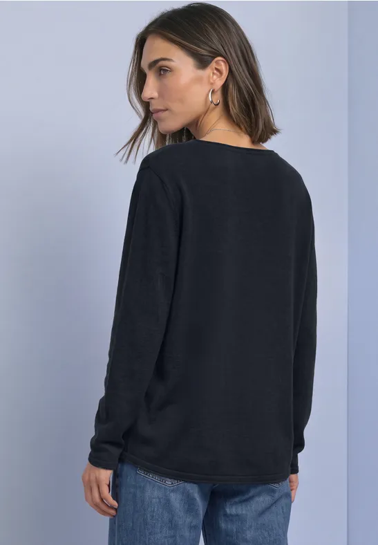 Frau in schwarzem Pullover und blauen Jeans, seitlich abgewandt vor hellem Hintergrund.