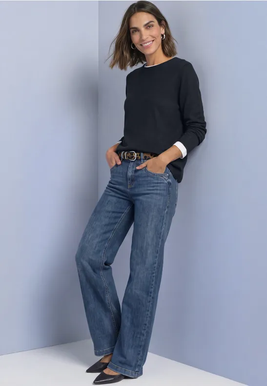 Frau in schwarzem Pullover und weit geschnittenen Jeans posiert lächelnd mit Händen in den Taschen.