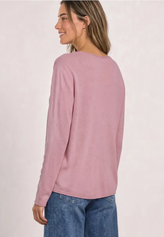 Eine Frau in rosa Langarmshirt und blauen Jeans steht seitlich, lächelt.