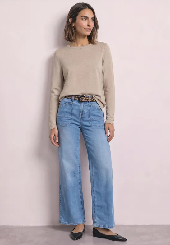 Frau in beigem Pullover, weiten hellblauen Jeans mit braunem Gürtel und schwarzen Schuhen vor hellem Hintergrund.