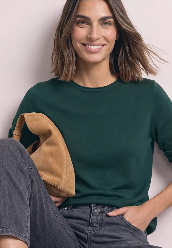 Frau sitzt lächelnd in dunkelgrünem Pullover, Jeans und hält eine braune Tasche.
