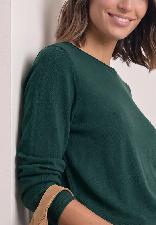 Eine Frau lehnt an einer Wand, trägt ein dunkelgrünes Langarmshirt und lächelt sanft.