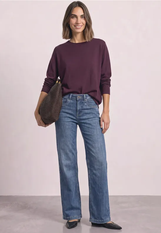 Een glimlachende vrouw in paarse trui, wijde jeans, witte sneakers en tas, staand voor een lichte achtergrond.