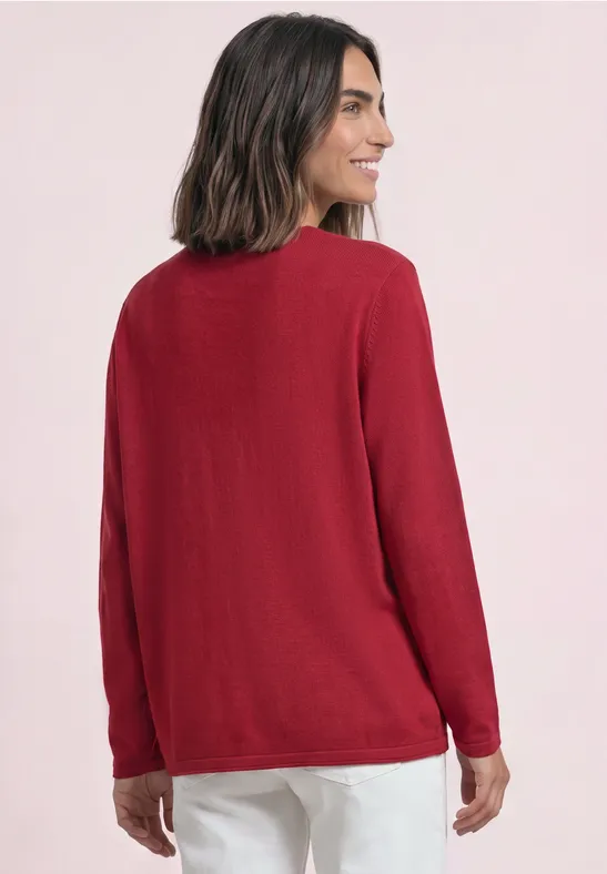 Rückansicht einer Frau in rotem Pullover und weißer Hose vor hellem Hintergrund.