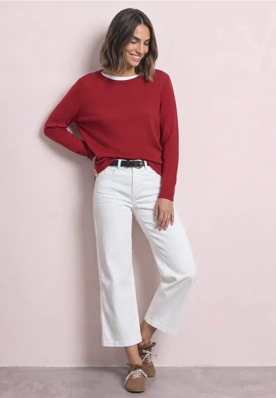 Frau in rotem Pullover, weißen Culotte-Jeans und braunen Sandalen vor rosa Hintergrund.