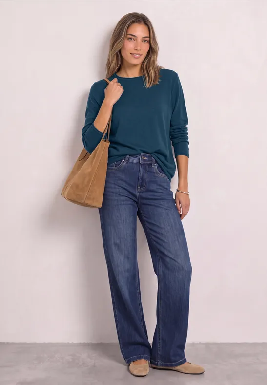Eine Frau trägt ein dunkelgrünes Langarmshirt, weite Jeans, eine braune Schultertasche und passende Schuhe.