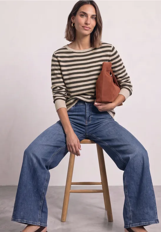 Frau sitzt auf Hocker, trägt gestreiften Pullover, weite Jeans und hält eine braune Tasche.