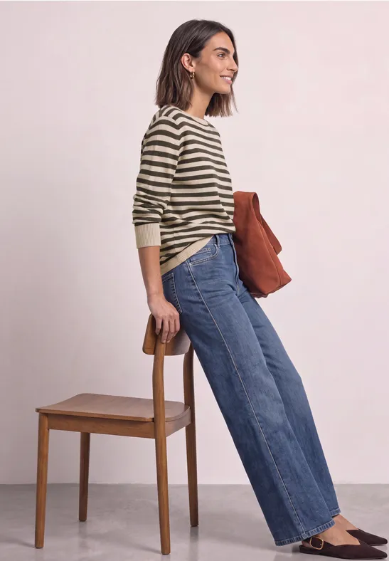 Frau in gestreiftem Pullover und weitem Jeans lehnt an Stuhl und hält eine braune Tasche.