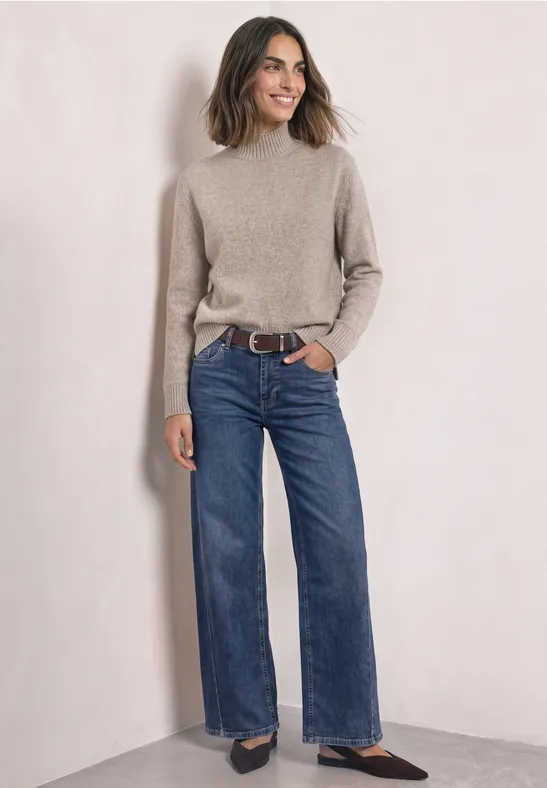 Femme debout devant un mur clair, portant un pull beige, un jean large et des chaussures foncées.