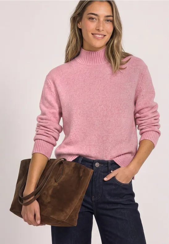 Frau mit rosa Pullover und dunklen Jeans hält eine braune Wildleder-Tasche und lächelt.