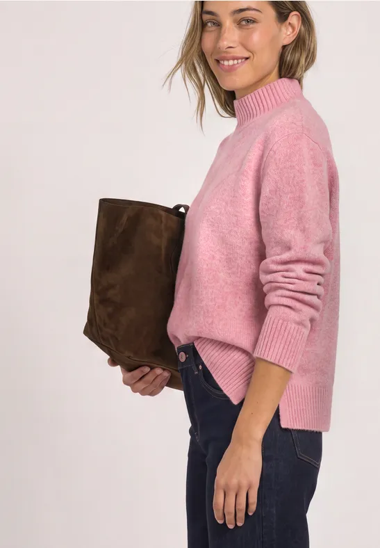 Frau in rosa Pullover und Jeans hält eine braune Tasche und lächelt seitlich.