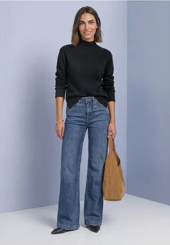 Frau in schwarzem Rollkragenpullover und weiten Jeans, hält eine braune Tasche vor schlichtem Hintergrund.