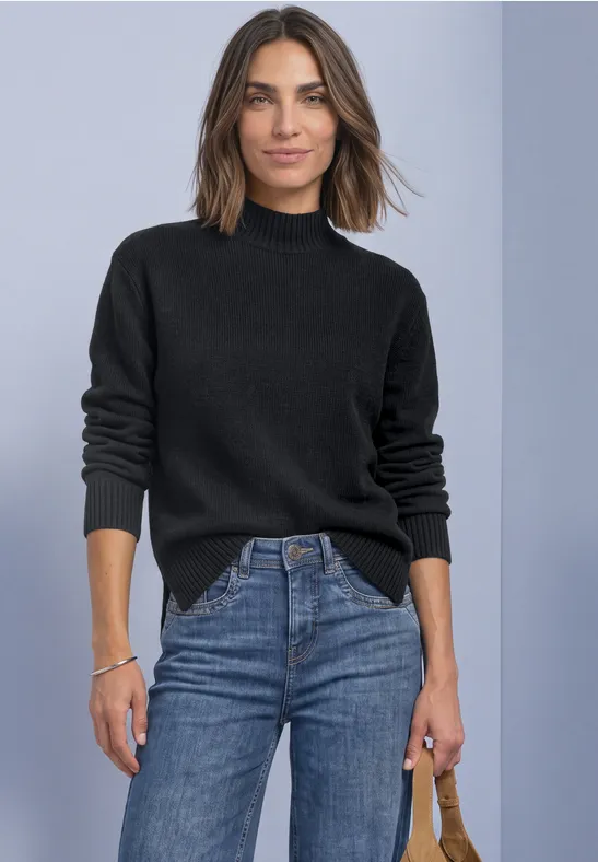 Frau in schwarzem Rollkragenpullover und blauen Jeans steht vor grauem Hintergrund, hält eine braune Tasche.