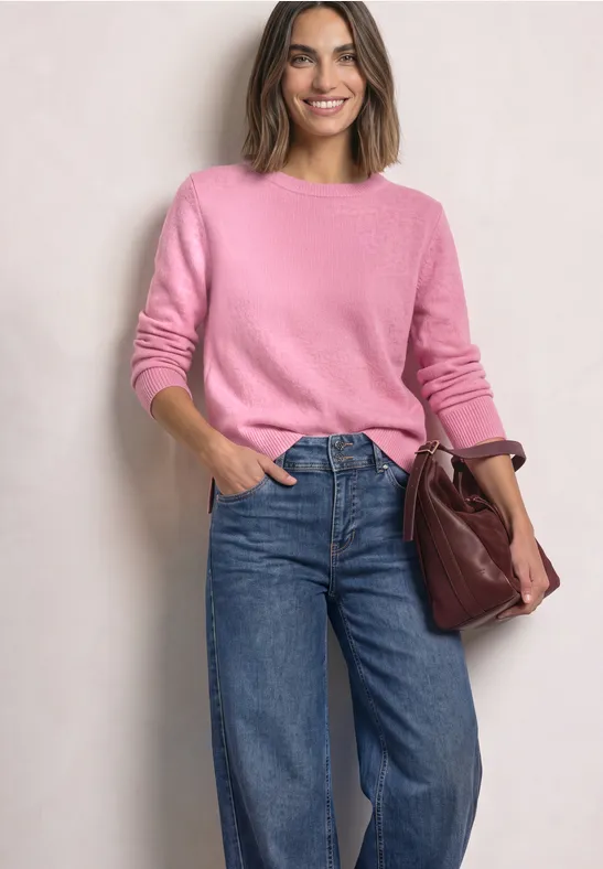 Eine Frau in rosa Pullover und Jeans lächelt und hält eine dunkelrote Tasche vor hellem Hintergrund.