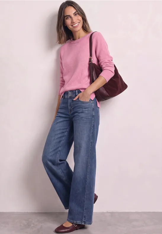 Junge Frau steht lächelnd in weitem Jeanshosen und rosa Pullover, mit brauner Schultertasche.